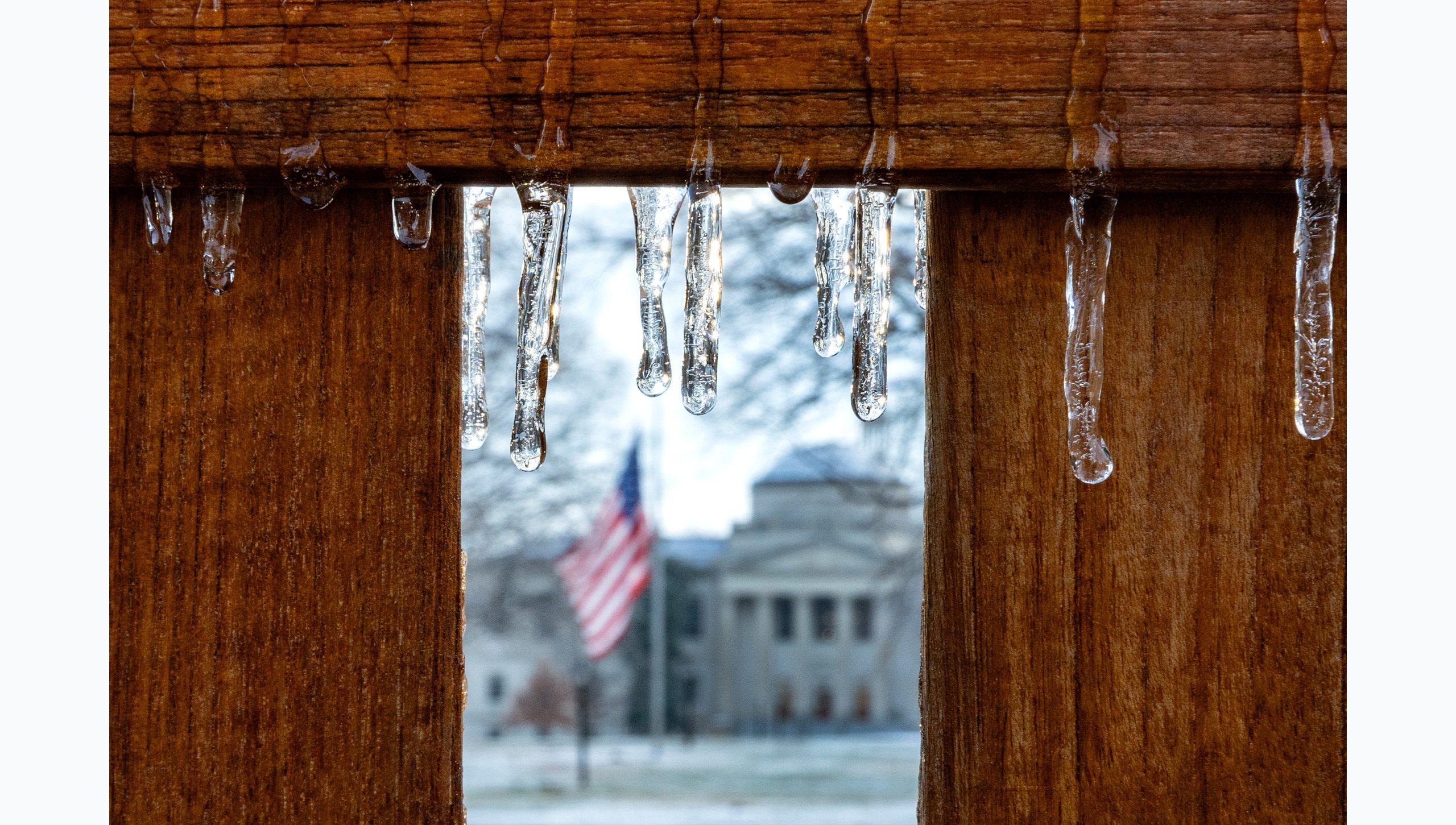 Close-up image of icicles hanging off a wooden bench on the campus of U.N.C Chapel Hill campus. Seen in the background is an American Flag and Wilson Library.